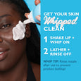Person applying white whipped cleanser to face from pump bottle against blue background.