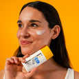 Woman holding white and yellow sunscreen bottle with white serum swatch applied to cheekbone against orange background.