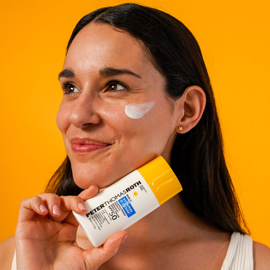 Woman holding white and yellow sunscreen bottle with white serum swatch applied to cheekbone against orange background.