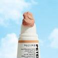 White pump bottle with beige tinted moisturizer swirl on top and tan stripe band, displayed against blue sky background.