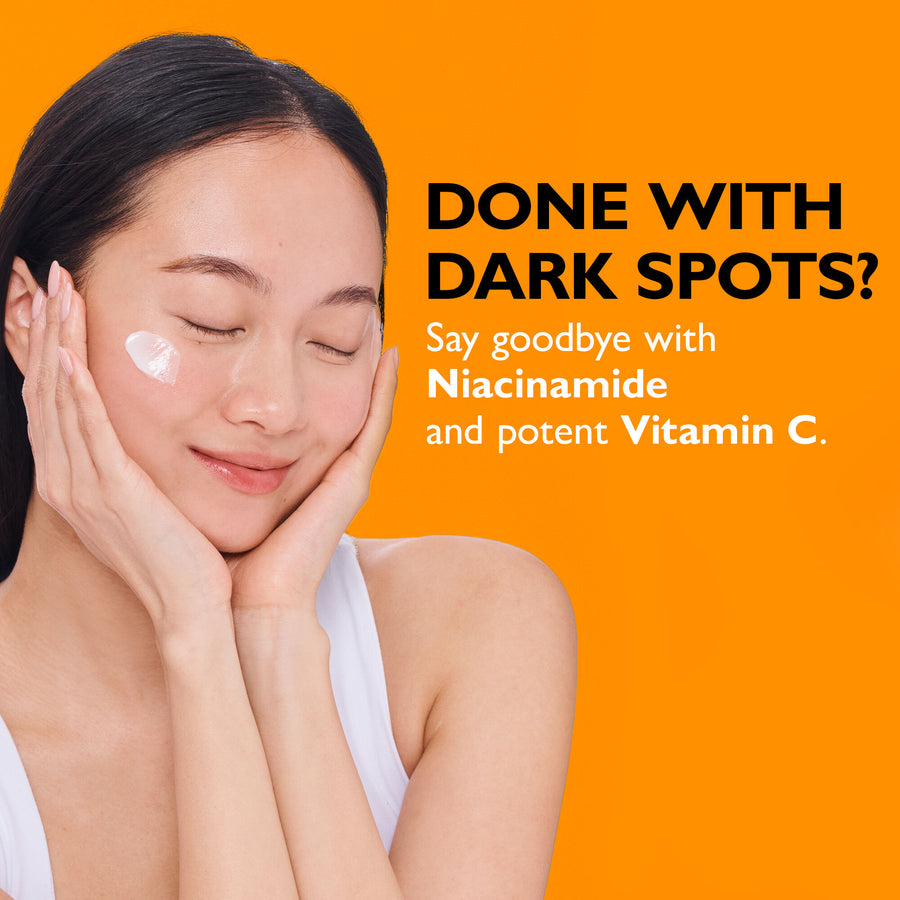 Woman with dark hair applying white cream to cheekbone against orange background.