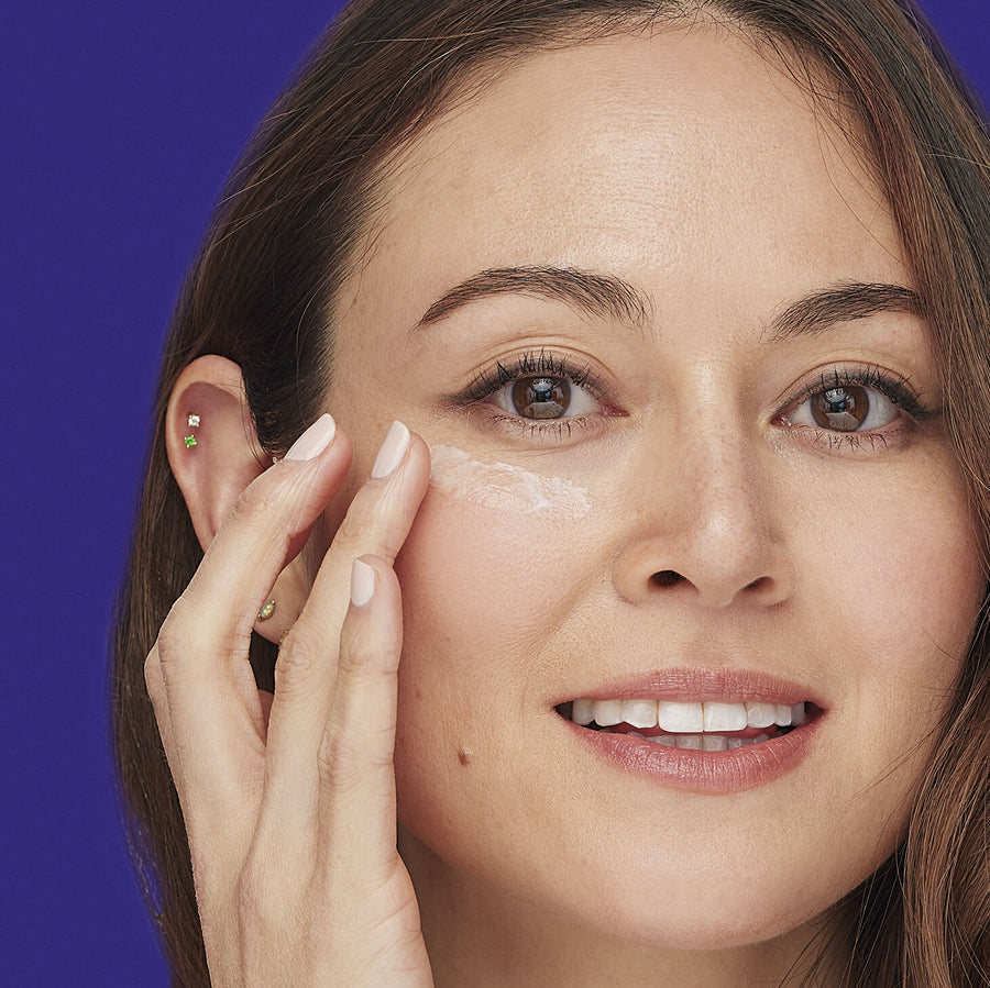 Woman applying cream to under-eye area with finger against purple background.