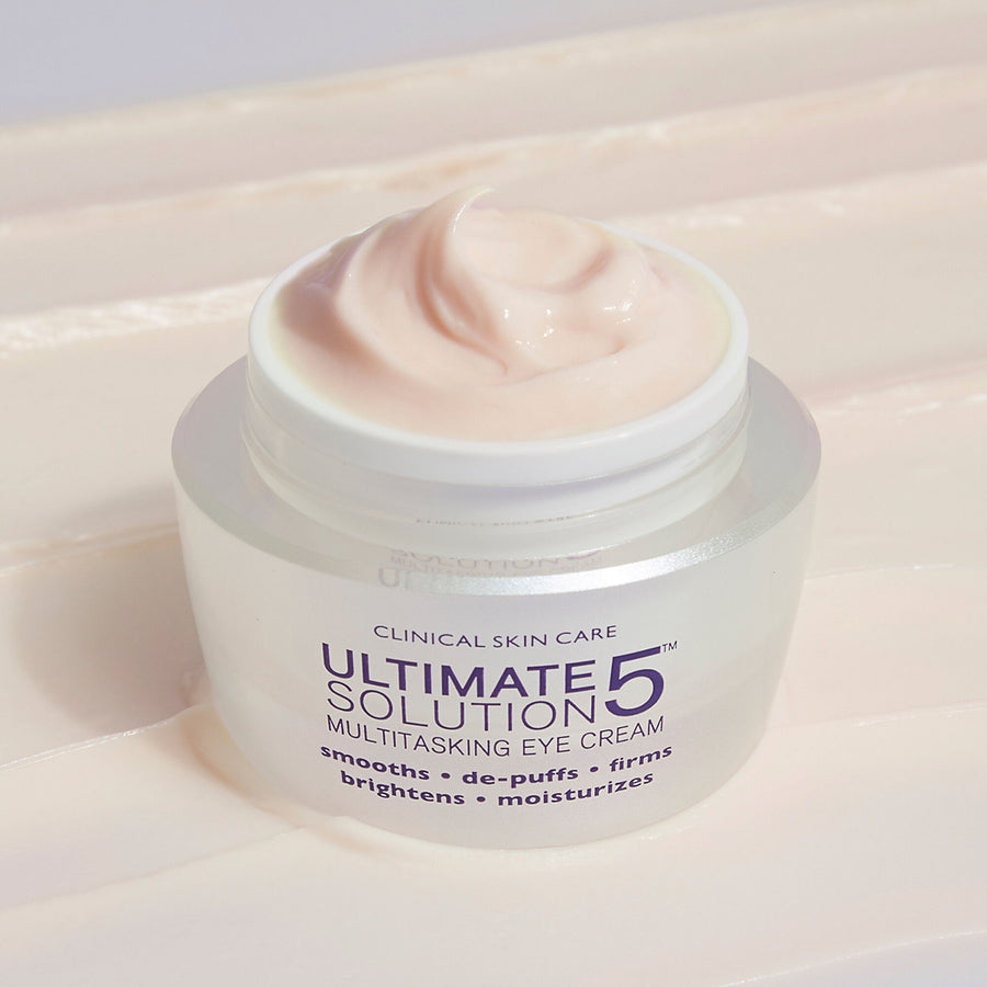 White jar of Ultimate Solution 5 Multitasking Eye Cream with pale pink cream visible in opened container.