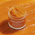 Orange pumpkin enzyme mask in white jar with textured surface displayed against pumpkin-colored background.